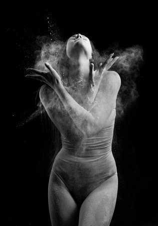 A beautiful slender ballet dancer girl wearing a bodysuit, sensually poses among the flying flour which covers her body, on a black background. Artistic, commercial, monochrome design.の写真素材