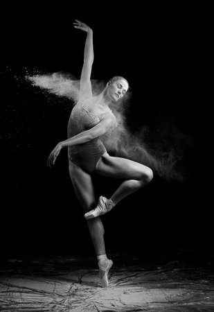 A beautiful slender ballet dancer girl wearing a bodysuit, sensually poses among the flying flour which covers her body, on a black background. Artistic, commercial, monochrome design.の写真素材