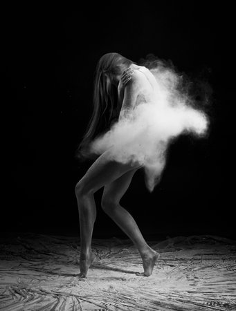 A beautiful slender ballet dancer girl wearing a bodysuit, sensually poses among the flying flour which covers her body, on a black background. Artistic, commercial, monochrome design.の写真素材
