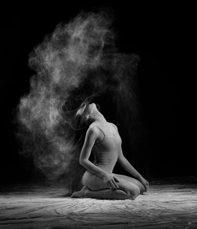 A beautiful slender ballet dancer girl wearing a bodysuit, sensually poses among the flying flour which covers her body, on a black background. Artistic, commercial, monochrome design.の写真素材