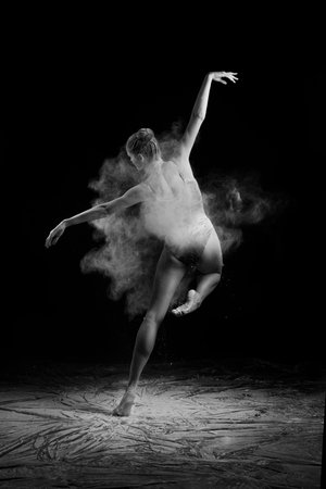 A beautiful slender ballet dancer girl wearing a bodysuit, sensually poses among the flying flour which covers her body, on a black background. Artistic, commercial, monochrome design.の写真素材