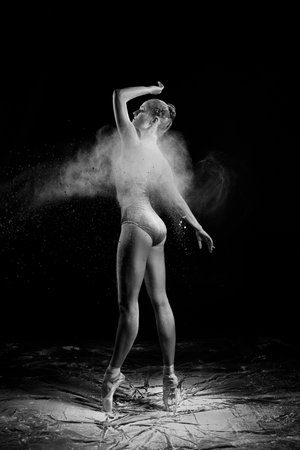 A beautiful slender ballet dancer girl wearing a bodysuit, sensually poses among the flying flour which covers her body, on a black background. Artistic, commercial, monochrome design.の写真素材