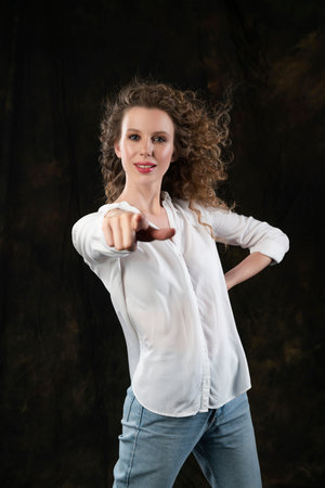 A beautiful girl, wearing jeans and a blouse, cheerfully points her finger to front, posing in the wind, which blows her long curly hair and clothes. Close up. Advertising, commercial, design.の写真素材