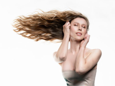 A beautiful girl with large breasts and naked shoulders sensually touches her face with hands, while her long hair flutters horizontally to the side. Isolated on white. Advertising, commercial design.の写真素材