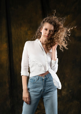 A beautiful girl, wearing jeans and a blouse, poses cheerfully in the wind, which blows her long curly hair and clothes. Advertising, commercial design.の写真素材