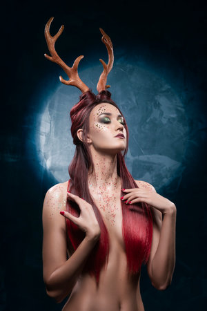 A beautiful sexy redhead topless girl, with deer horns on her head and sequins on her body, sensually posing, covering her naked breasts with her long hair. Conceptual, commercial, christmas design.の写真素材
