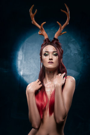 A beautiful sexy redhead topless girl, with deer horns on her head and sequins on her body, sensually posing, covering her naked breasts with her long hair. Conceptual, commercial, christmas design.の写真素材