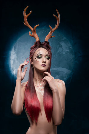 A beautiful sexy redhead topless girl, with deer horns on her head and sequins on her body, sensually posing, covering her naked breasts with her long hair. Conceptual, commercial, christmas design.の写真素材