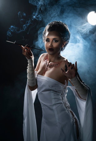 Beautiful girl wearing a Halloween costume with rich make-up and an imitation of an autopsy seam on her chest is emotionally posing holding a cigarette holder in the smoke. Vintage, cosplay design.の写真素材