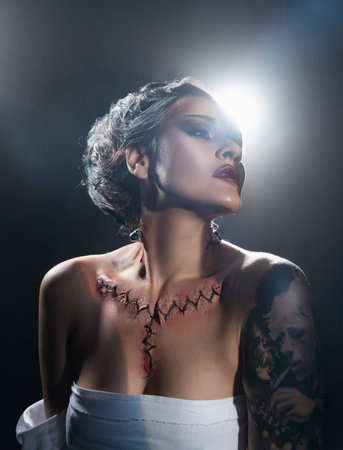 Beautiful girl wearing a Halloween costume with rich make-up and an imitation of an autopsy seam on her chest is emotionally posing in the smoke. Vintage, cosplay, artistic design.の写真素材