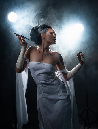 Beautiful girl wearing a Halloween costume with rich make-up and an imitation of an autopsy seam on her chest is emotionally posing holding a cigarette holder in the smoke. Vintage, cosplay design.の写真素材