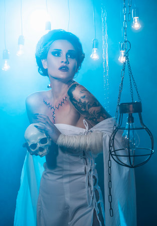 Girl wearing a Halloween costume with rich make-up and an imitation of an autopsy seam on her chest is emotionally posing in decorations holding a skull in her hands. Holiday, artistic design.の写真素材