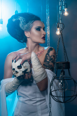 Girl wearing a Halloween costume with rich make-up and an imitation of an autopsy seam on her chest is emotionally posing in decorations holding a skull in her hands. Holiday, artistic design.の写真素材