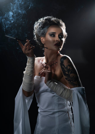 Beautiful girl wearing a Halloween costume with rich make-up and an imitation of an autopsy seam on her chest is emotionally posing holding a cigarette holder in her hand. Vintage, cosplay design.の写真素材