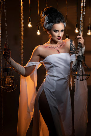 A beautiful girl wearing a Halloween carnival costume with rich make-up and an imitation of an autopsy seam on her chest is emotionally posing in decorations. Artistic, holiday, commercial design.の写真素材