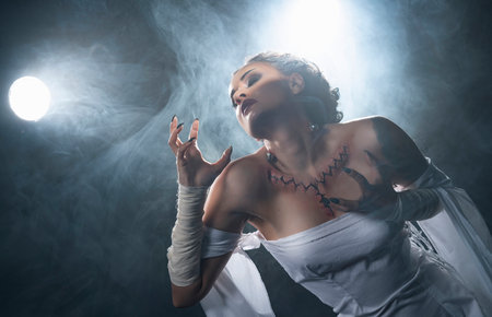 Beautiful girl wearing a Halloween costume with rich make-up and an imitation of an autopsy seam on her chest is emotionally posing in the smoke. Vintage, cosplay, artistic design.の写真素材