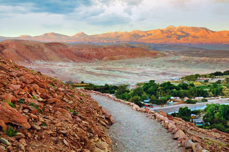 View of the desert and village near the archeological site of Pukara de Quitor, surrounded by red rocky hills and green vegetation against a blue sky covered by clouds.の写真素材