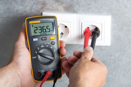 Electrician checking voltage in socket with tester. Professional multimeter in hand close-up.