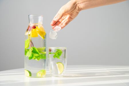 Summer refreshing drink. Water with lemon, lime and mint. Woman hand drops ice cubes into glass with...
