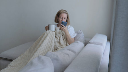 A young woman drinks hot tea while lying on the sofa. A woman on the sofa, covered with a blanket,...