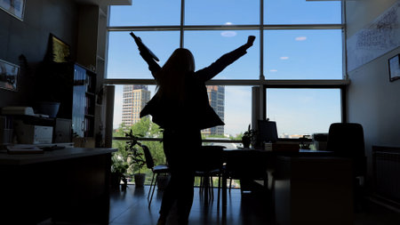 Silhouette of person with raised arms in office moment of triumph and success career achievement. Person silhouette with arms raised high in office signifies victorious moment of success career.の写真素材