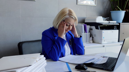 Tired businesswoman holds head sitting at desk overworking emotional stress burnout mental health. Caucasian businesswoman in office visibly stressed holding head overwork burnout emotional strain.の写真素材