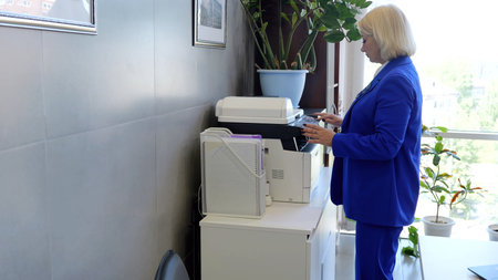 Millennial caucasian businesswoman in suit stands near copy machine in office making copies of...