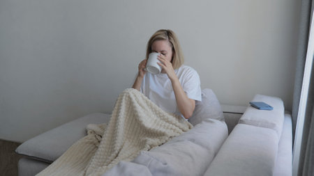 Young woman covered with a warm blanket drinks tea at home. A young woman drinks hot tea while lying on the sofa. A woman on the sofa, covered with a blanket, drinks a hot drink.の写真素材