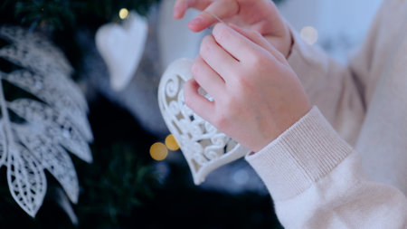 Woman happily adorns a christmas tree with white decorations, encircled by glowing lights, setting a warm and festive ambiance, getting ready for the holiday season with love and cheerの写真素材