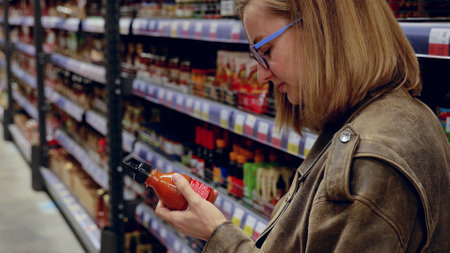 Customer with glasses reading label on hot sauce bottle while shopping for groceries in supermarket aisleの写真素材