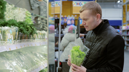Man choosing greens in supermarket, shelf with greens. Man making healthy food choices in a supermarket, carefully selecting fresh produce among a variety of optionsの写真素材