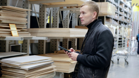 Customer in hardware store uses smartphone to compare wooden boards in lumber aisle. A man in a hardware store, choosing lumber, shelves with lumber.の写真素材