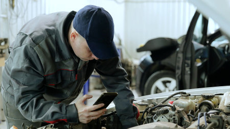 Experienced automotive technician checking smartphone while diagnosing car engine problems in modern repair workshop, focusing on precise mechanical diagnosticsの写真素材