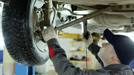 Skilled mechanic wearing protective gear examining vehicle undercarriage, using flashlight to check suspension details while car positioned on lifting hoistの写真素材
