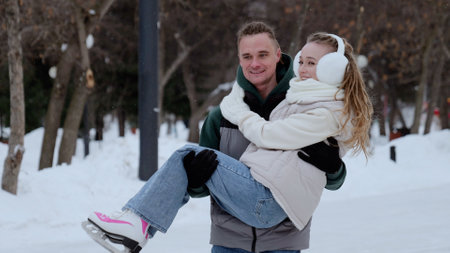 Cheerful man lifting woman wearing ice skates, sharing romantic moment amid snow covered park landscape, expressing winter happiness through playful connectionの写真素材