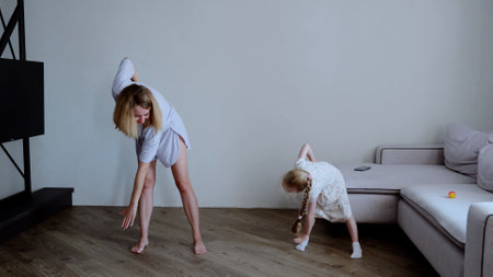 Mother and daughter in casual attire, doing a forward bend and stretching exercises together in their homeの写真素材