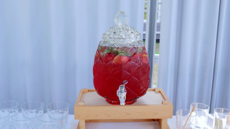 Glass beverage dispenser holding vibrant red fruit punch, garnishing with ice, mint, fresh fruit...
