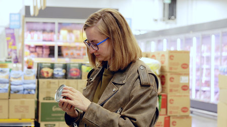Modern health conscious shopper, a young woman reads a can label in a supermarket aisle, considering...