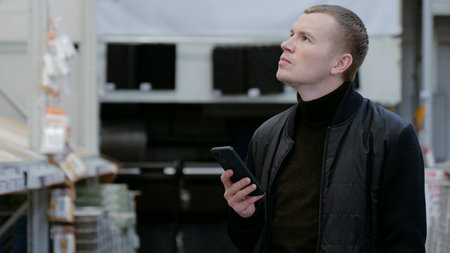 Contractor browsing building materials on a smartphone in a lumber yard or home improvement store. A man in a hardware store, choosing flooring, shelves with ceramic tiles.の写真素材