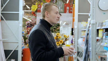 Man carefully choosing light bulb in hardware store, comparing options for home improvement project. Focus on energy efficiency and product detailsの写真素材