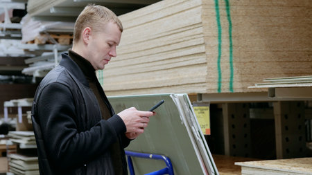 A man in a hardware store, choosing lumber, shelves with lumber. Customer uses a smartphone to compare prices, check availability, and find information on construction materials at a hardware storeの写真素材