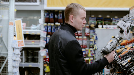 Construction worker comparing cordless drill features and prices while shopping in a hardware store, examining design and specifications. A man in a hardware store choosing a tool.の写真素材