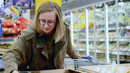 Attentive woman shopping for groceries in supermarket, inspecting products by refrigerated vegetable display, showcasing mindful consumer habitsの写真素材