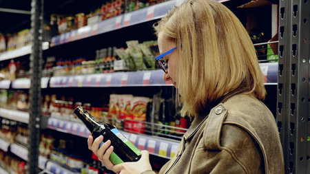 Customer examines label on dark glass bottle while shopping for beverages in grocery store, considering ingredients and price before making purchaseの写真素材