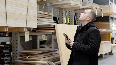 Carpenter checking wooden boards, selecting quality materials using smartphone, standing amid stacked timber and plywood in warehouse settingの写真素材