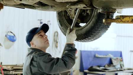 Skilled mechanic fixing vehicle suspension with wrench, working intently under raised car in...
