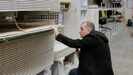 Construction worker checking drywall sheets at hardware store, comparing sizes and quality for renovation project, selecting precise building materials with professional attentionの写真素材