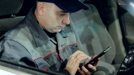 Professional mechanic reviewing smartphone inside darkened car, scrolling automotive resources, managing repair details during late evening workshop hoursの写真素材