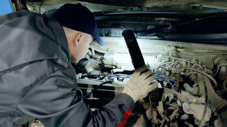 Experienced mechanic wearing protective gloves examining car engine, using flashlight while performing detailed diagnostic work inside dimly lit automotive workshopの写真素材