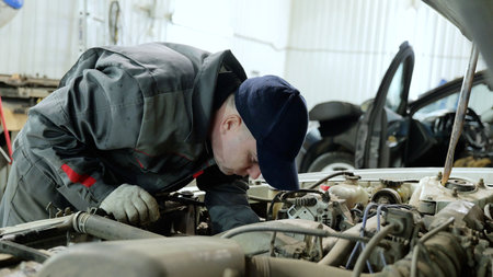 Skilled mechanic checking car engine, wearing protective gear in well lit automotive workshop, focusing on detailed technical maintenance and repair workの写真素材